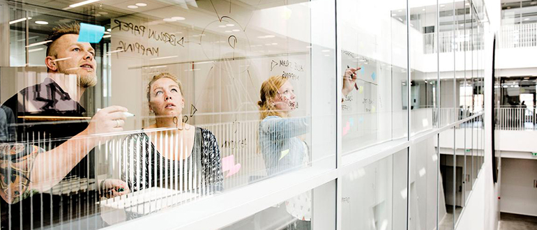 Studerende samarbejder ved glasvæg på The Faculty of Engineering - SDU Tre personer samarbejder ved en glasvæg i en moderne bygning og skriver på glasset.
