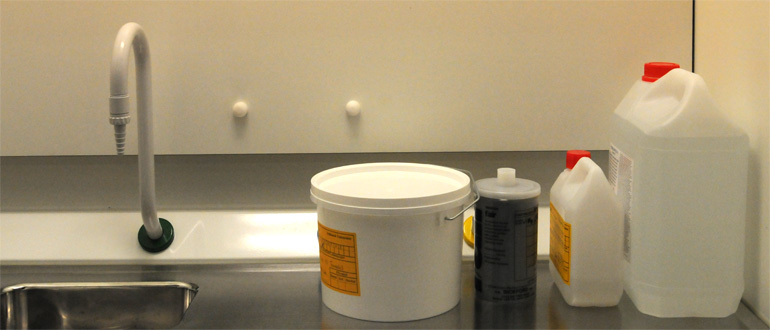Laboratory sink with a white bucket and multiple chemical containers on a countertop.