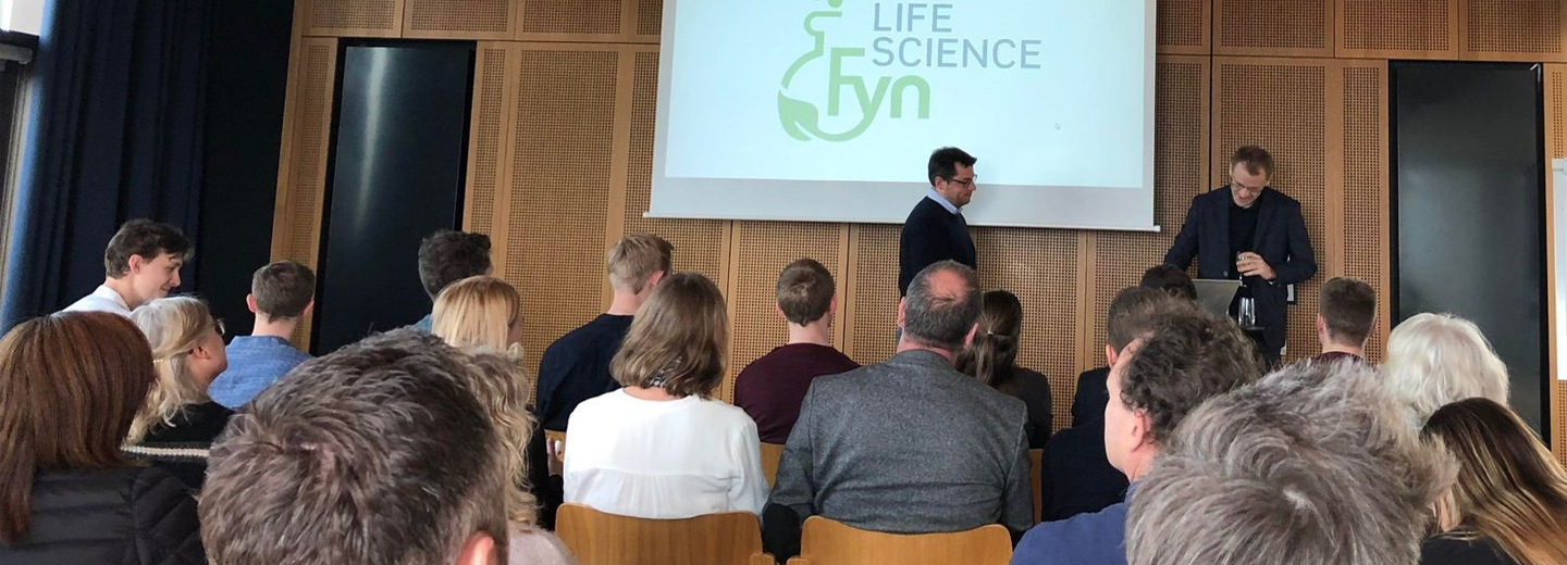 Publikum i et mødelokale foran to oplægsholdere og en skærm med “LIFE SCIENCE fyn”.