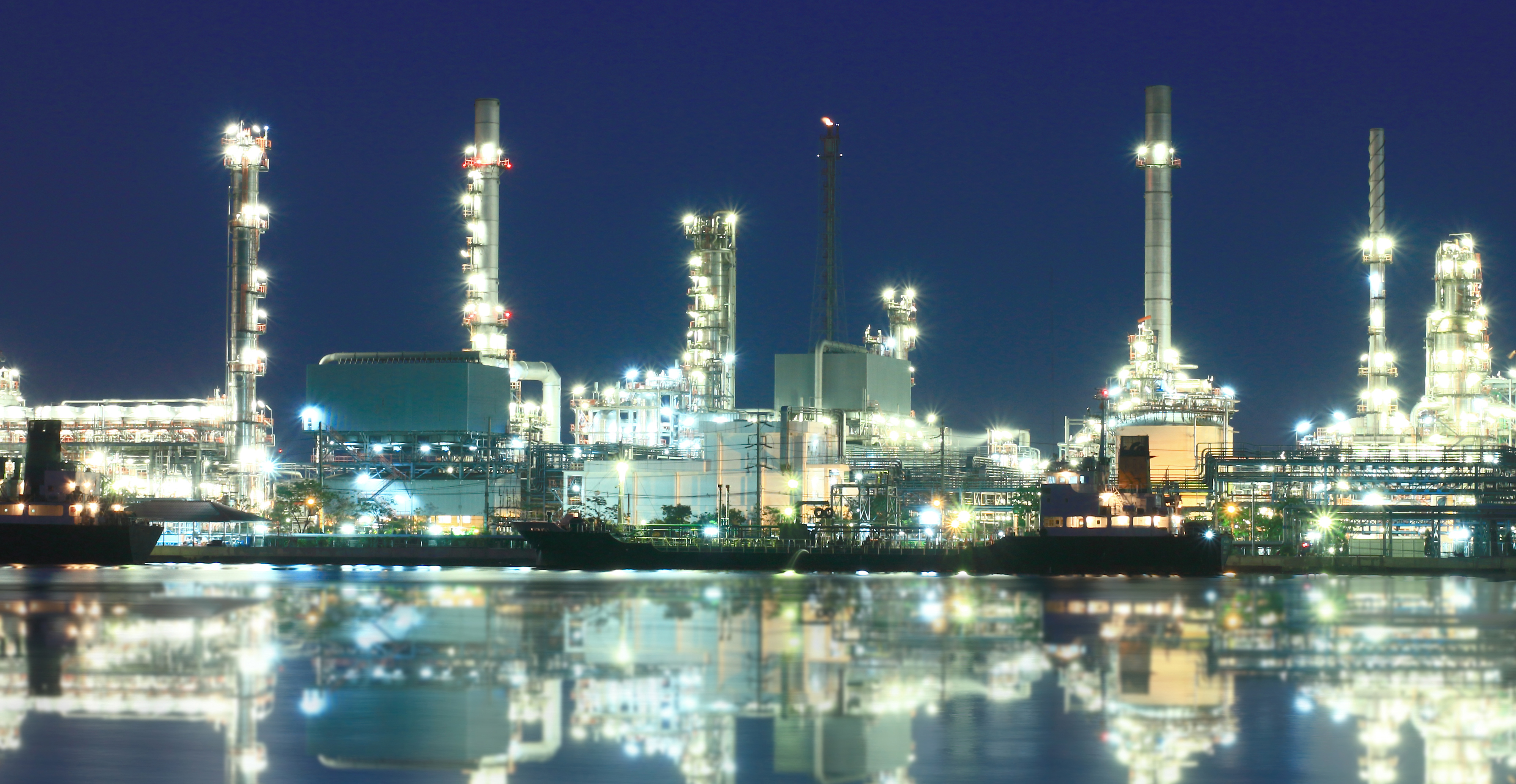 Oplyst industriraffinaderi om natten ved vand med skibe og refleksioner.