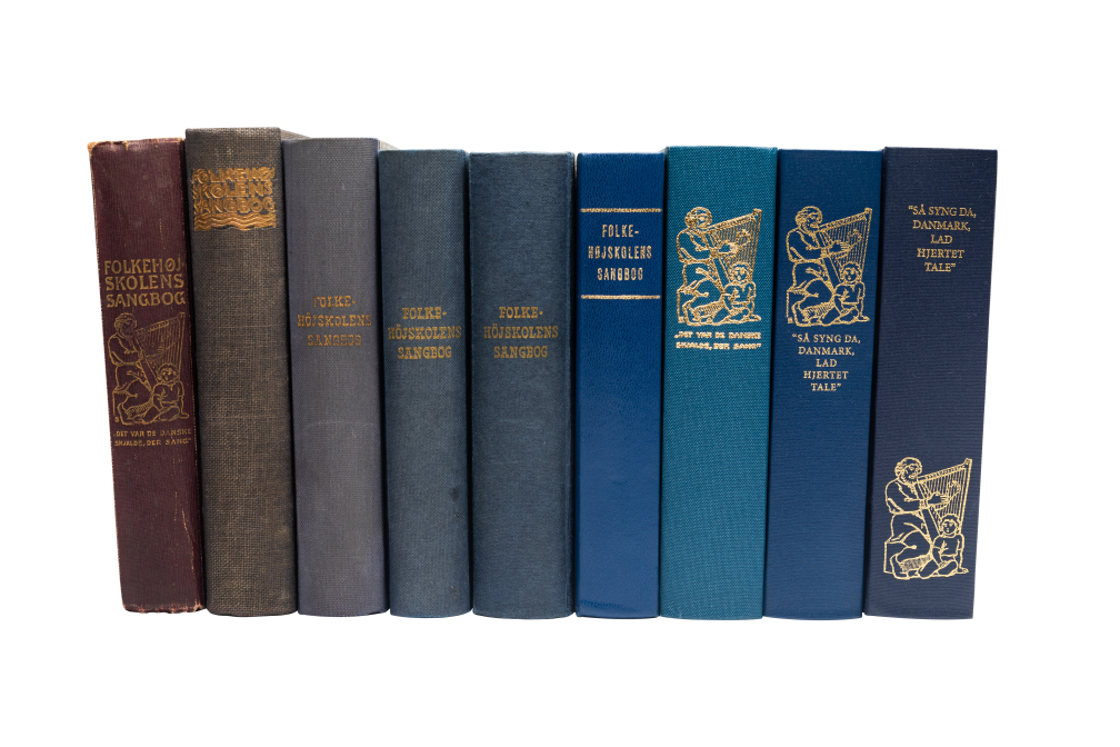 En række gamle udgaver af højskolesangbøger stående på række. Bøgerne har forskellige farver og titler, herunder 'Folkehøjskolens sangbog' og 'Så syng da, Danmark, lad hjertet tale'. 