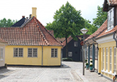 Gule historiske bygninger med røde tegltage omkring et brostensbelagt gårdrum og en sort port i baggrunden.