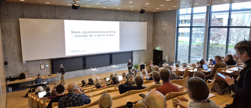 Billede fra Center for Grundskoleforsknings årlige konference. Billedet viser salen fyldt med mennesker og peger op mod scenen, hvor man kan se en powerpoint-præsentation med et oplæg