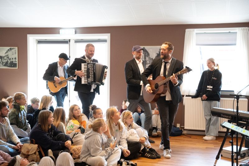 Musikere spiller for en gruppe skoleelever, der sidder på gulvet i et lokale, mens en Roland-keyboardstation står til højre.