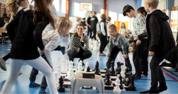 Børn og lærer der samles om et spil skoleskak i idrætssalen.