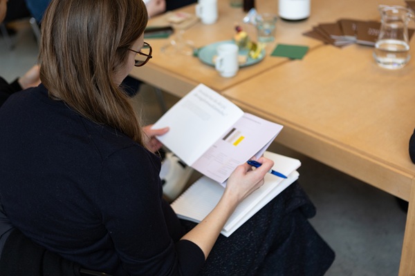 Alt text: A person is sitting at a table reading a report with graphs and text while taking notes in a notebook. On the table are cups, water carafes, and papers, suggesting a meeting or workshop. The image relates to Center for upper secondary school research at SDU.
