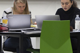 To kvinder ved et bord med bærbare computere og en grøn stol i forgrunden.