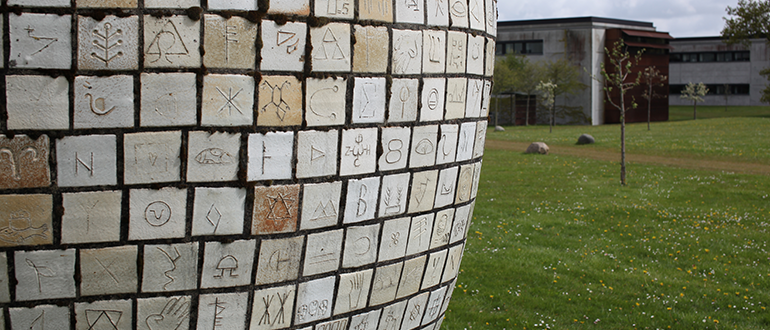 Buet skulptur med mange indgraverede fliser foran en græsplæne og bygninger.