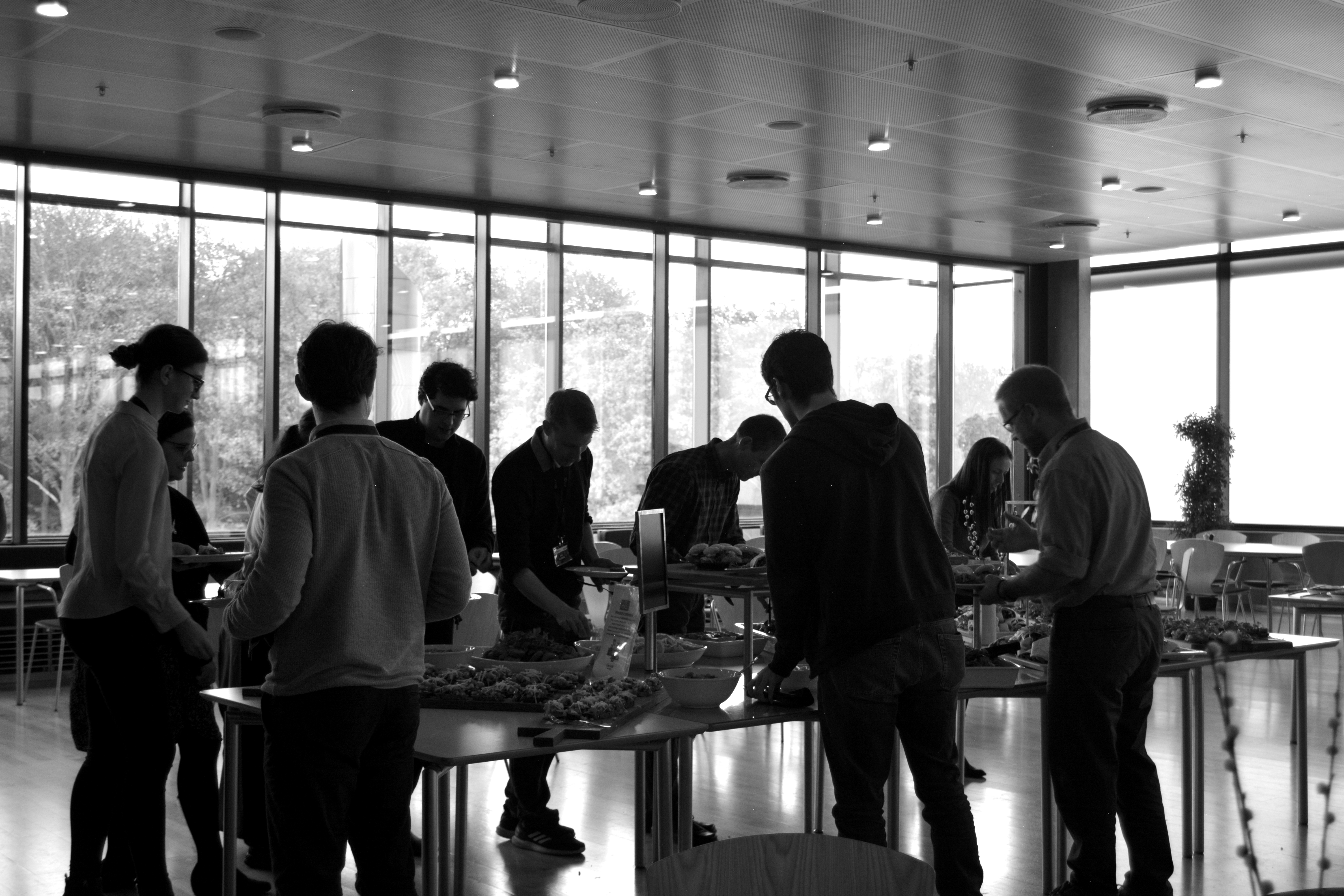 Several people standing around buffet tables at an indoor event in a bright room with large windows, shown in black and white.
