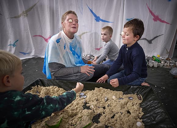 Stine Mariegaard og en dreng, sidder ved sandkassen; begge med en muslingeskal foran det ene øje som en monokel. 