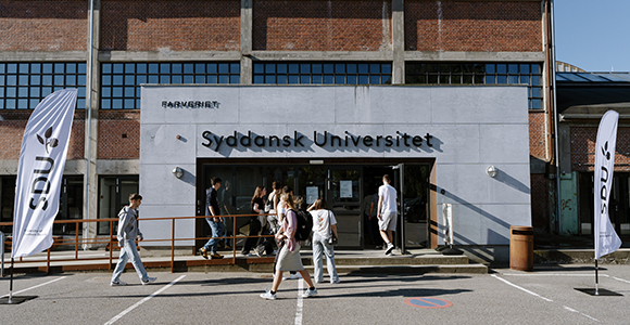 Studerende går foran indgangen til Syddansk Universitet, flankeret af to hvide "SDU"-flag.