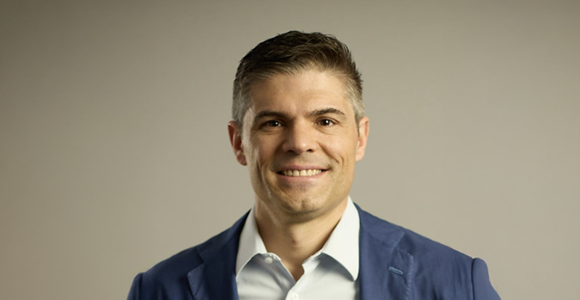 Smiling man in a blue blazer and light-colored shirt in a studio portrait against a neutral background.
