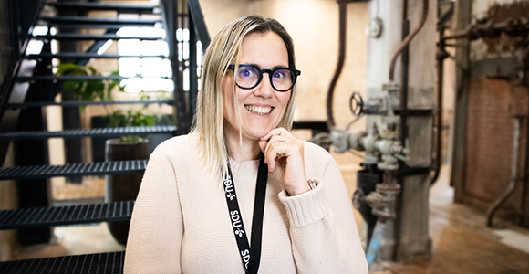 Smiling woman with glasses in a light sweater standing indoors near a staircase and industrial equipment.