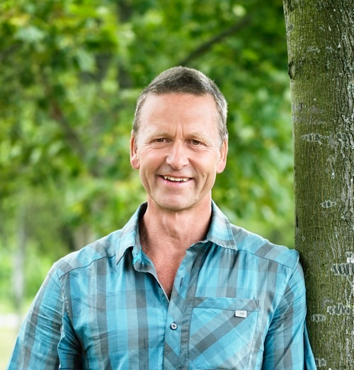 Smilende mand i blåternet skjorte står udendørs ved siden af et træ.