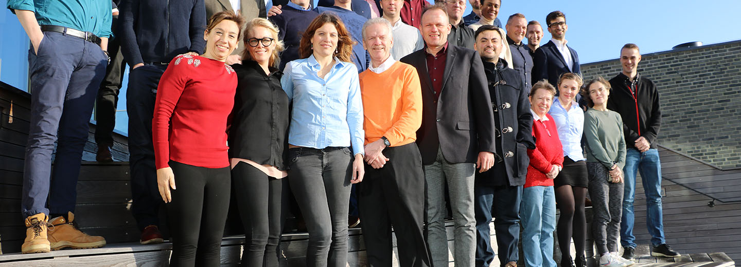 Large group of adults posing together on outdoor steps outside a building in bright sunlight.