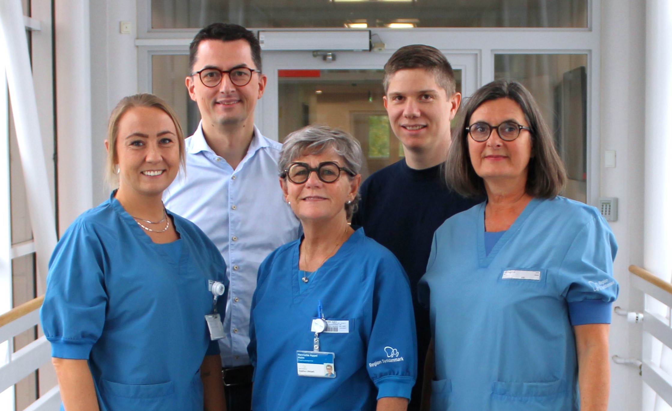 Fem personer poser i en hospitals- eller klinikgang, hvor tre er i blå arbejdsdragter og to er i almindeligt tøj. Teksten "Region Sjælland" er synlig på en uniform.