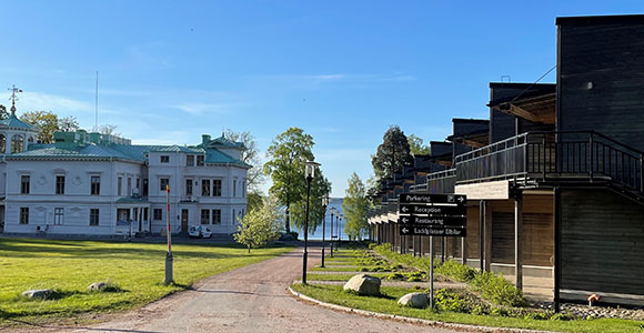 Scenic view of a gravel path between a historic white manor house and modern wooden buildings with balconies. Signposts indicate directions to parking, reception, and restaurant. Lush green lawns, trees, and a glimpse of the water in the background highlight the picturesque location under a clear blue sky.