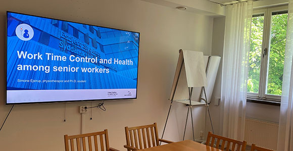 Meeting room with a large screen displaying a presentation titled "Work Time Control and Health among senior workers" by Simone Ejstrup, physiotherapist and PhD student. The room features a large window with a view of green trees, a flip chart on a stand, and wooden chairs around a conference table.
