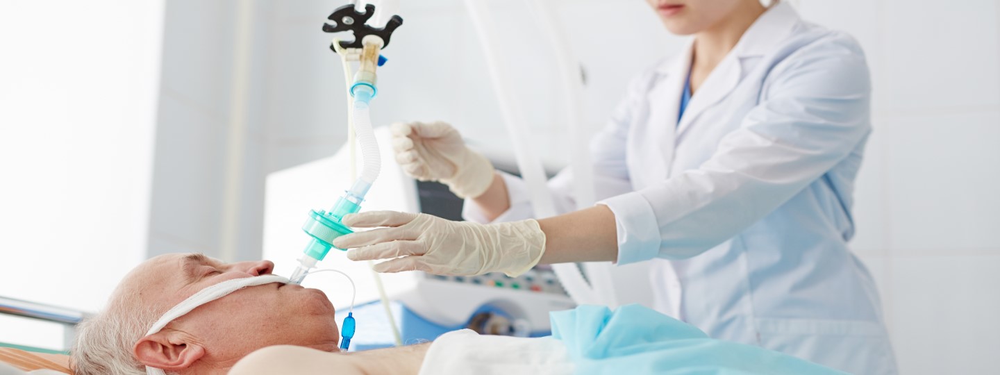 Ældre patient i hospitalseng med endotrakealtube, mens en sundhedsperson justerer respiratorudstyr.