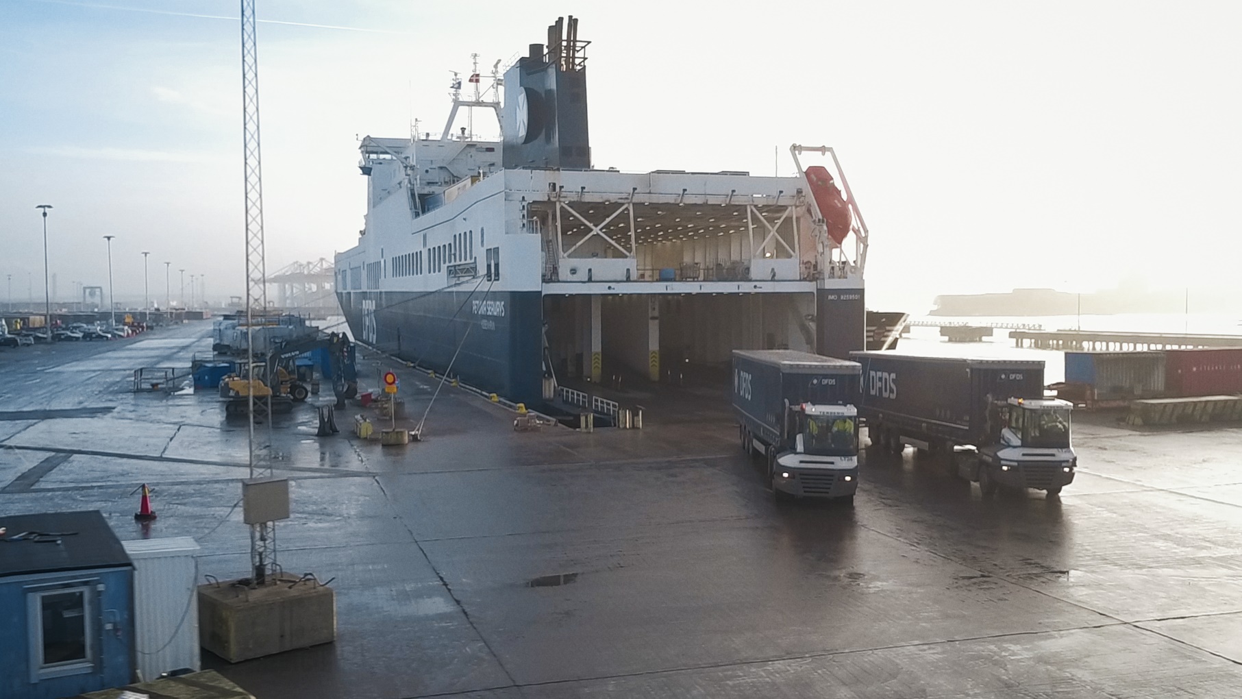 DFDS ferry at a port with the rear loading ramp open and two trucks with "DFDS" trailers on the wet dockside.