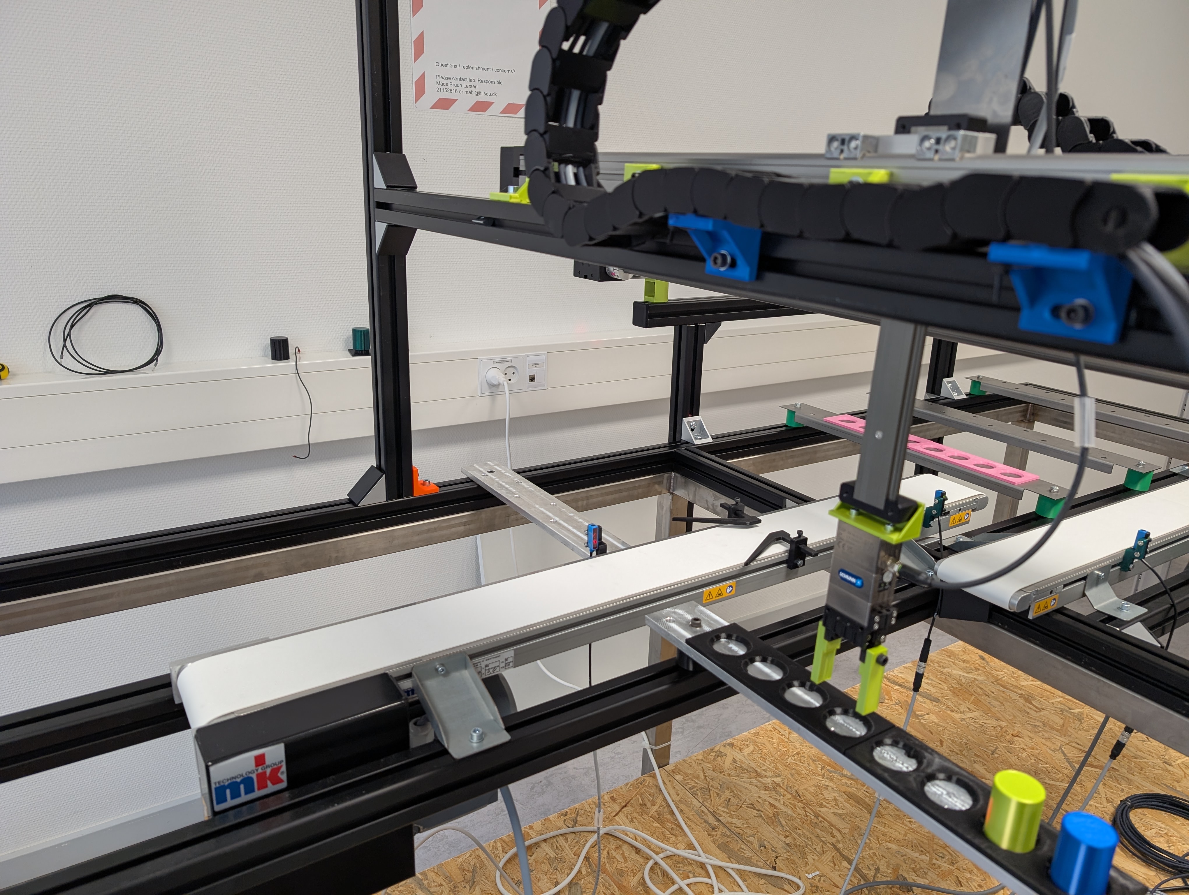 Industrial automation machine with linear rails, cable chain, robotic gripper, fixture with circular parts, and an "mk" logo on the front component.
