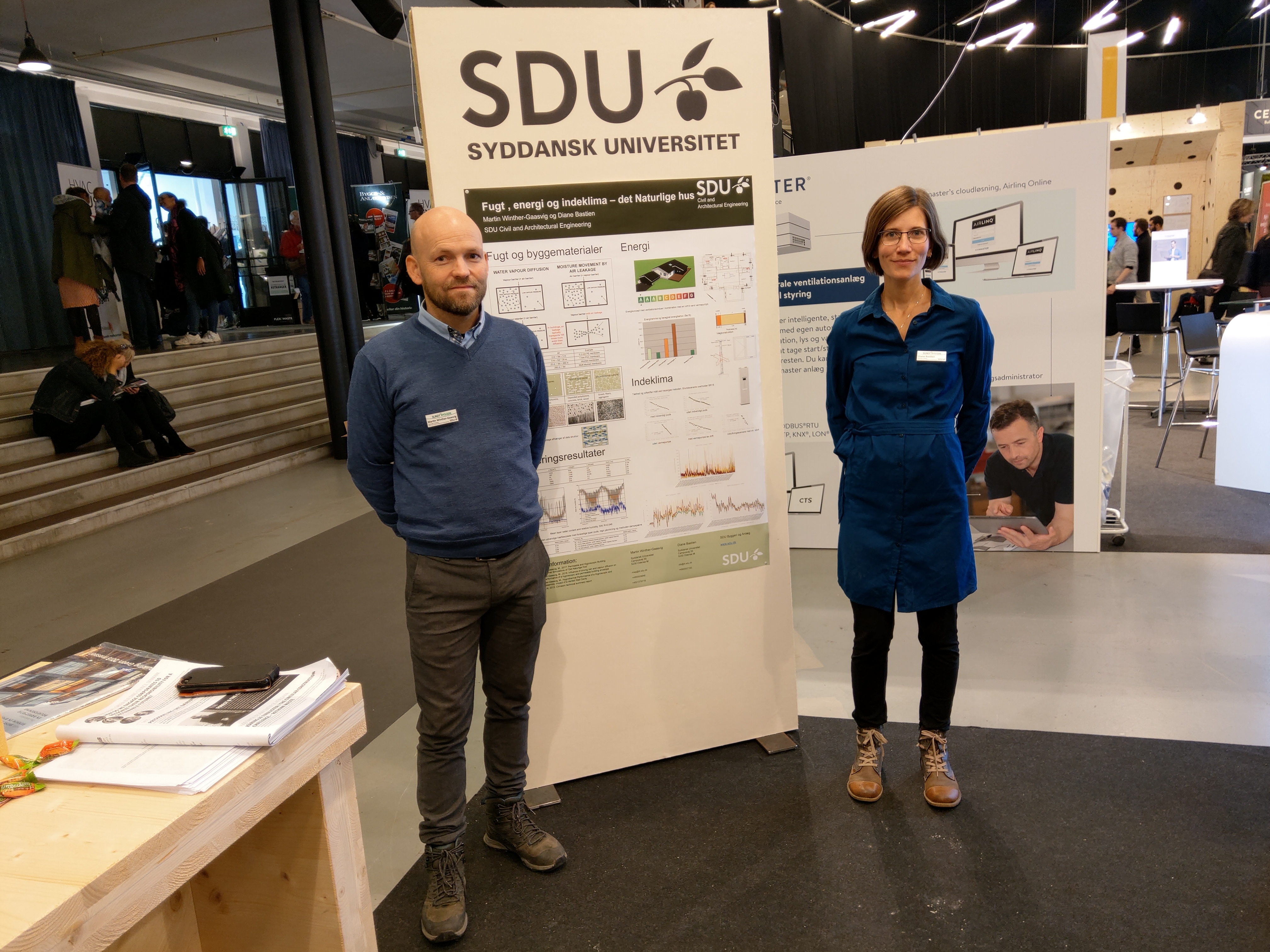 To personer står foran en SDU-udstillingsstand med plakater og teksten "SDU" og "SYDDANSK UNIVERSITET".