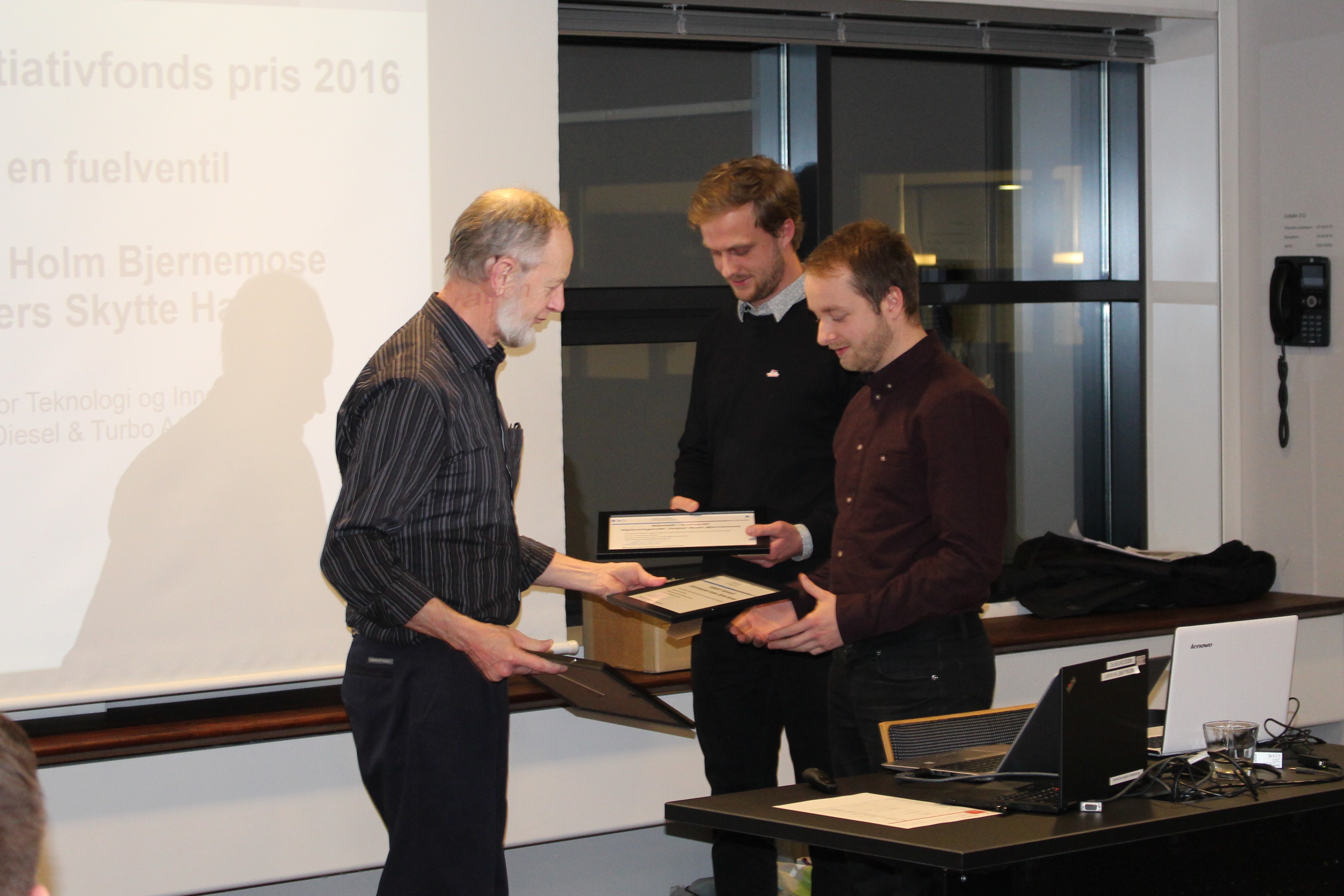 Tre mænd udveksler diplomer eller plaketter foran et lærred med delvis synlig tekst, herunder "tiativfonds pris 2016".
