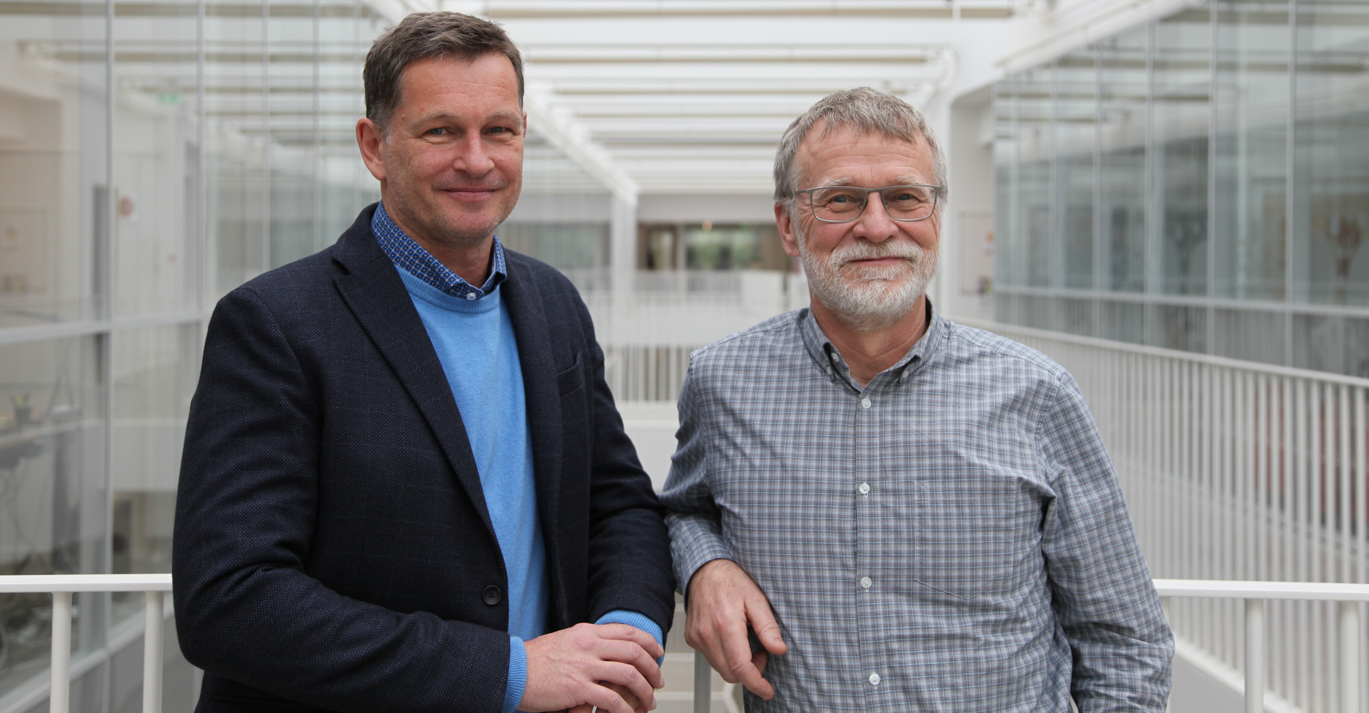 To smilende mænd står i et lyst atrium med glasvægge og hvide gelændere; billedet er knyttet til Jan Vang Peter Hasle.