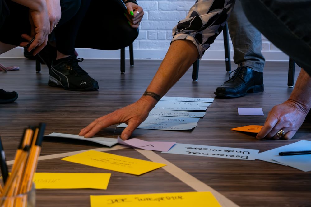 Personer i en workshop sidder og knæler omkring farvede kort og papirark på gulvet.