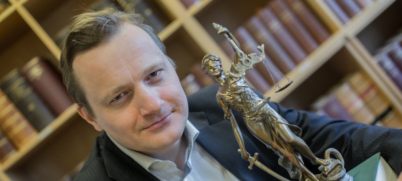 Mand ved siden af en Justitia-statuette foran bogreoler med bøger.