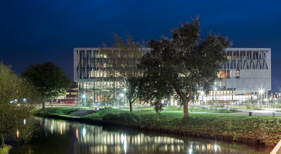 Oplyst moderne bygning ved en kanal om natten med træer foran og refleksioner i vandet.