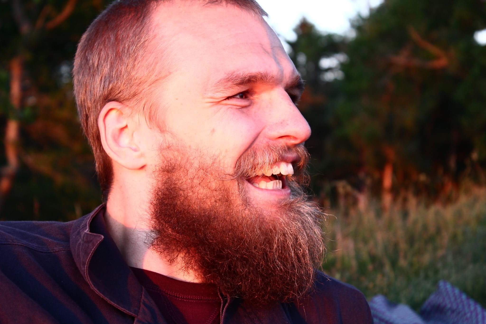 Smiling bearded man in side profile outdoors at sunset with trees and grass behind him.