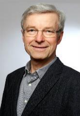 Older man with gray hair and glasses in a dark blazer, photographed against a light gray studio background.