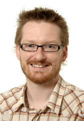 Portræt af Kasper Klein Portræt af en smilende person med briller, lyst hår og skæg mod en lys baggrund.