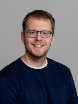 Benjamin Bakke Hansen Portrætfoto af en smilende mand med kort brunt hår, skægstubbe og briller, iført en mørkeblå sweatshirt, fotograferet foran en neutral grå baggrund.