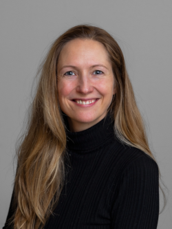 Portrætfoto af en smilende kvinde med langt lyst hår, iført en sort rullekravetrøje, fotograferet foran en neutral grå baggrund.