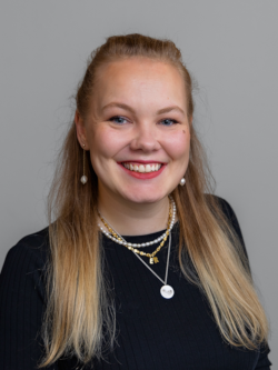 Portrætfoto af en smilende kvinde med langt blondt hår, iført en sort bluse og flere halskæder, fotograferet foran en neutral grå baggrund.