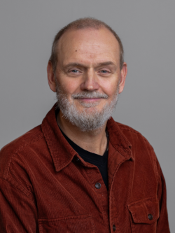 Lars Morsø Portrætfoto af en smilende mand med kort gråt hår og gråt skæg, iført en rustrød fløjlsskjorte over en sort T-shirt, fotograferet foran en neutral grå baggrund.