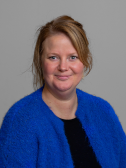 Portrait photo of a smiling woman with light reddish-blonde hair, wearing a blue jacket over a black blouse, photographed against a neutral grey background.