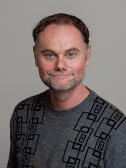 Portrætfoto af en mand med kort mørkt hår og let skægstub, iført en grå striktrøje med sort mønster, fotograferet foran en neutral grå baggrund.