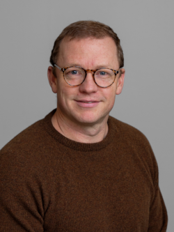 Portrætfoto af en mand med kort lyst hår og briller, iført en brun striktrøje, fotograferet foran en neutral grå baggrund.