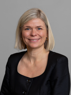 Portrætfoto af en smilende kvinde med kort blondt hår, iført en sort bluse, fotograferet foran en neutral grå baggrund.