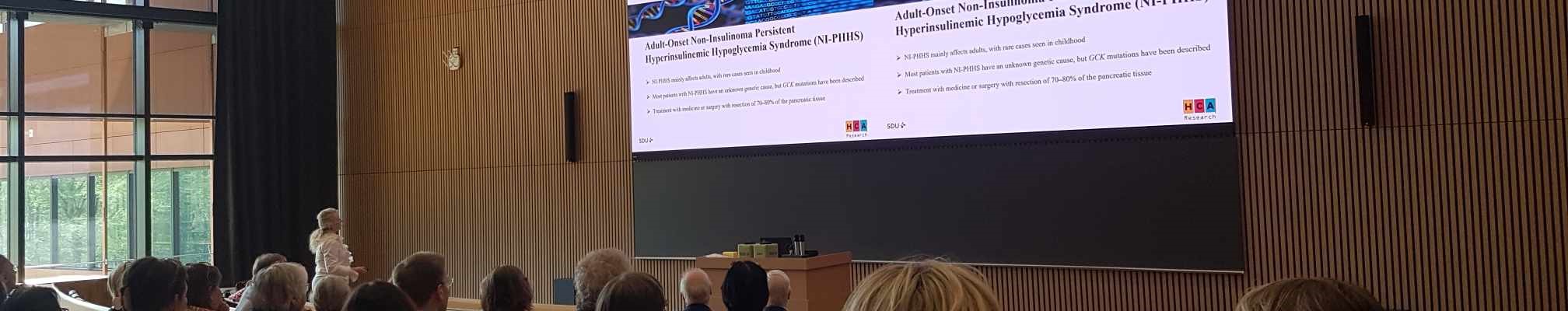 Oplægsholder foran et publikum i et auditorium med medicinske slides projiceret på en stor skærm.