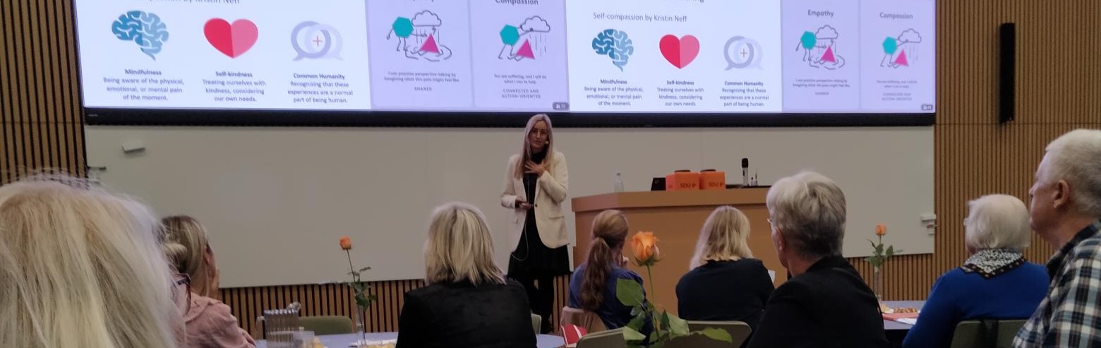 Kvinde taler foran et siddende publikum i et auditorium med slides om “Mindfulness”, “Self-kindness”, “Common Humanity”, “Empathy” og “Compassion” på en storskærm.
