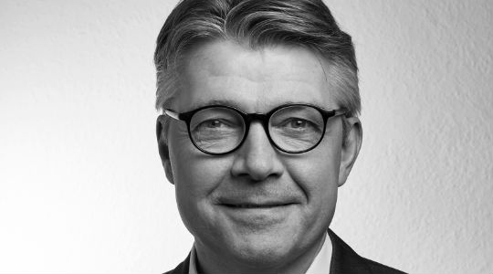 Black-and-white headshot of a man wearing round glasses and a suit jacket.