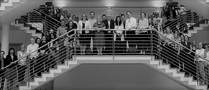 Stor gruppe på trappe i kontor- eller institutionsmiljø Stor gruppe mennesker poser på en bred indendørs dobbelttrappe i et sort-hvidt foto.