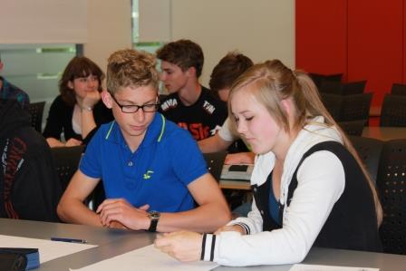 Two students seated at a table reviewing papers, with other students in the background in a classroom or workshop.