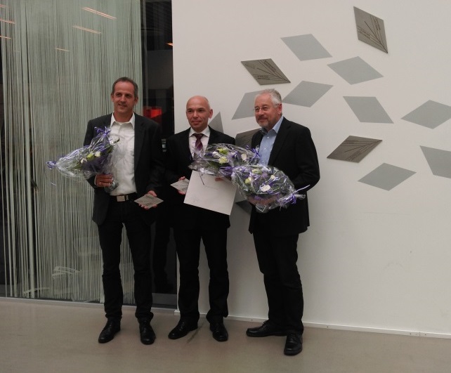 Three men in suits holding flower bouquets and documents while posing indoors by a decorated wall.