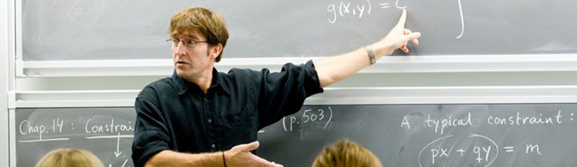Underviser foran tavle peger på matematiske formler og tekst som "g(x,y)=c" og "px + qy = m".