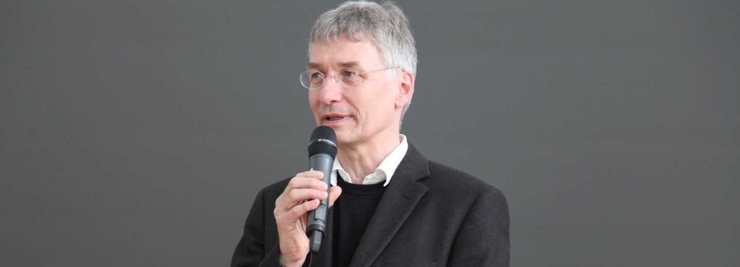Horst Gnter Rubahn holder en mikrofon og taler foran en grå baggrund.